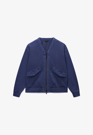 Navy blue zip-up jacket in soft fabric, featuring two front patch pockets, ribbed cuffs, and a v-neck design. Simple and functional.
