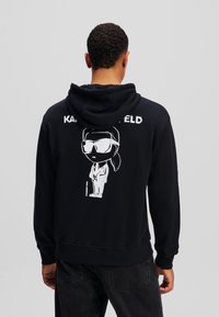 Sweatshirt negru cu glugă reglabilă, având pe spate un grafic de desene animate alb, care înfățișează un personaj cu ochelari de soare și costum.