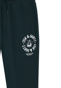 Donkergroene sweatpants met een trekkoord in de taille, met een witte grafische afbeelding van een schip en de tekst: "Gemaakt voor de dappere."