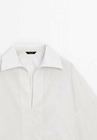 Chemise blanche à col avec une ouverture en V et manches courtes visible sur un fond blanc uni.