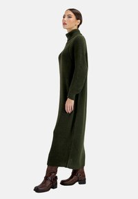 Langes, geripptes, dunkelgrünes Strickkleid mit hohem Kragen und langen Ärmeln, kombiniert mit braunen Stiefeletten mit Schnallenakzenten.