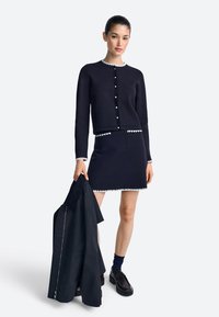 Tmavomodrý kardigán a sukňový set s bielymi zubatými okrajmi. Kardigán má gombíky na prednej strane. Modelky držia zodpovedajúci tmavý kabát.