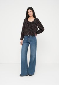 Μαύρο φλοράλ πουκάμισο με τετράγωνο ντεκολτέ και λεπτομέρεια φιόγκου, σε συνδυασμό με φαρδιά μπλε τζιν παντελόνια. Απλό φόντο.