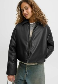 Czarna skórzana kurtka typu cropped z przednim zamkiem, szerokim kołnierzem i lekko bufkami, zestawiona z szarym swetrem i jasnoniebieskimi dżinsami.