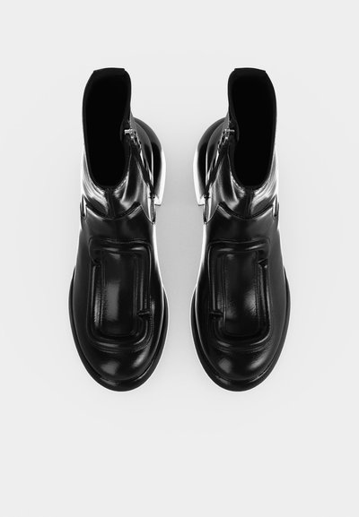 Paire de bottines en cuir noir brillant avec bouts arrondis, semelles épaisses et fermetures éclair, vues de dessus sur un fond clair.