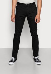 Pantaloni chino in cotone nero con vestibilità slim, frontale piatto e tasche laterali. Abbinati a sneaker alte nere e una camicia grigia chiara.
