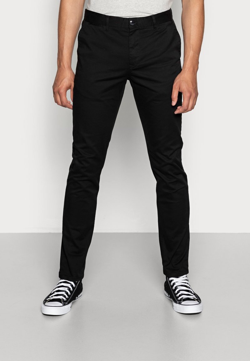 Pantaloni chino in cotone nero con vestibilità slim, frontale piatto e tasche laterali. Abbinati a sneaker alte nere e una camicia grigia chiara.