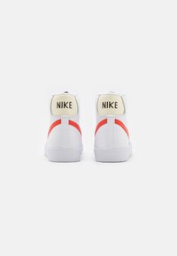 Sneakers bianchi alta con una striscia di accento rossa, tacco rotondo e logo Nike nero in contrasto su una toppa beige sul retro.
