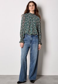 Grüne, blumenmusterte Bluse mit langen Ärmeln und Rüschensäumen, kombiniert mit hochgeschnittenen, weit geschnittenen blauen Denim-Jeans. Schwarze Knöchelstiefel.