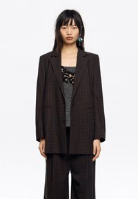 Blazer de cuadros marrón oversize con líneas azules, cierre de un solo botón y solapas con muesca. Lleva puesto sobre una blusa gris con detalles de encaje.