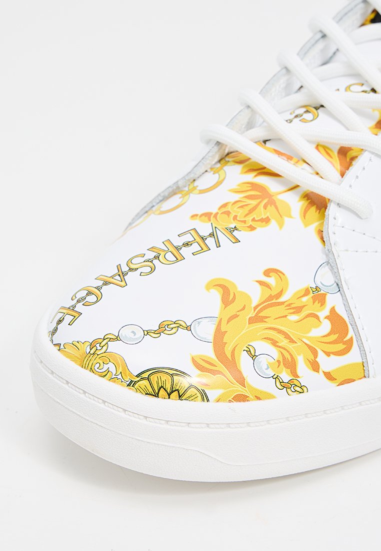 Baskets blanches avec un motif floral et chaînes dans des tons jaunes et dorés, avec un système de laçage et une texture lisse. Marquage visible.