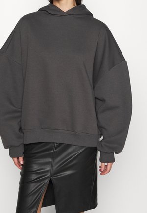 Kvinde iført oversized mørkegrå hættetrøje og sort lædernederdel med frontsplit, stående mod en ensfarvet hvid baggrund.