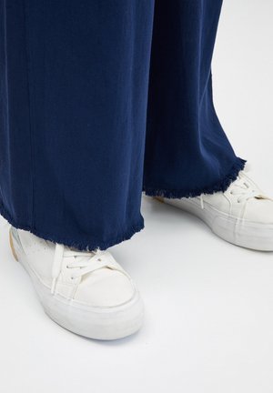 Námornícke nohavice s širokými nohavicami a roztrhnutými lemami, v kombinácii s bielymi teniskami s textúrovanou gumovou podrážkou a šnúrkami.