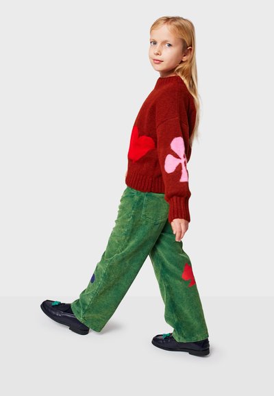 Hnedý pletený sveter s červenými a ružovými tvarmi, v kombinácii so zelenými kordovými nohavicami so vzormi v modrej a červenej farbe, a čiernymi topánkami s ozdobami.