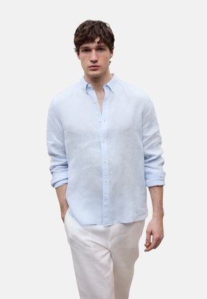 Junger Mann mit zerzaustem braunem Haar, trägt ein hellblaues Hemd mit hochgekrempelten Ärmeln und weiße Hose, steht vor einem einfarbigen Hintergrund.