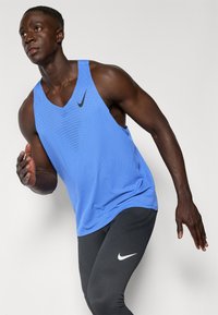 Niebieski sportowy tank top z perforowaną fakturą, z czarnym logo Nike Swoosh. W komplecie z czarnymi legginsami o fakturze, również z logo Nike.