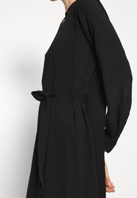Schwarzes Kleid mit langen Ärmeln, einem Bindegürtel, vertikaler Nadelstreifenstruktur und einem Schlüsselloch-Ausschnitt. Weicher Stoff mit fließendem Design.