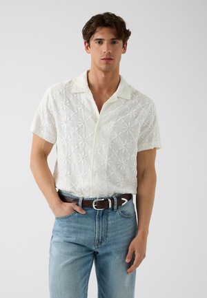 Jeune homme aux cheveux foncés portant une chemise blanche à manches courtes avec broderie florale et un jean bleu clair avec une ceinture marron.