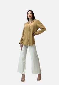 Camicia button-up beige chiaro con un tessuto testurizzato, abbinata a pantaloni bianchi a gamba larga e tacchi beige. La camicia ha un colletto e maniche lunghe.