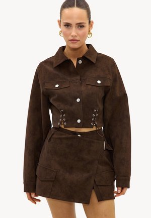 Femme portant une veste courte marron avec des côtés lacés et une jupe asymétrique assortie avec poches et détail boucle.