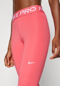 Roze sportlegging met een brede tailleband met de tekst "NIKE PRO", een gladde stof en een klein wit Nike-logo op de bovenbeen.