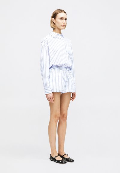 Γυναίκα που στέκεται φορώντας πουκάμισο με μακριά μανίκια με κάθετες ριγέ σε ανοιχτό μπλε και λευκό και ταιριαστό σορτς, με μαύρα φλατ παπούτσια διακοσμημένα με ασημένιες τρύπες.