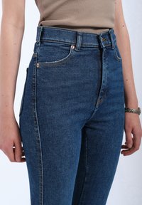 High-waisted blue denim jeans with copper rivets, contrasting stitching, and a fitted silhouette. Smooth texture with side pockets.