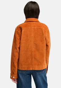 Orange kordflöjlsjacka med krage, långa ärmar och två knappade manschetter, med vertikal ribbad textur och rak skärning.