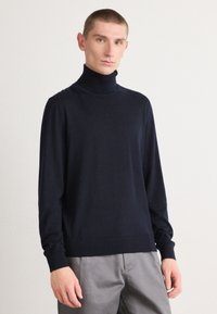 Sweater de gola alta azul-marinho em tecido suave, com mangas longas, silhueta ajustada e punhos canelados, combinado com calças cinza-claro.