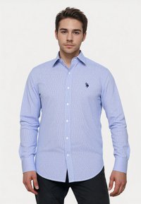 Giovane uomo che indossa una camicia a maniche lunghe a righe blu chiaro e bianche e pantaloni scuri, in piedi contro uno sfondo semplice.