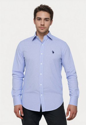 Joven vestido con una camisa de botones de rayas azul claro y blanca, de mangas largas, y pantalones oscuros, de pie contra un fondo liso.