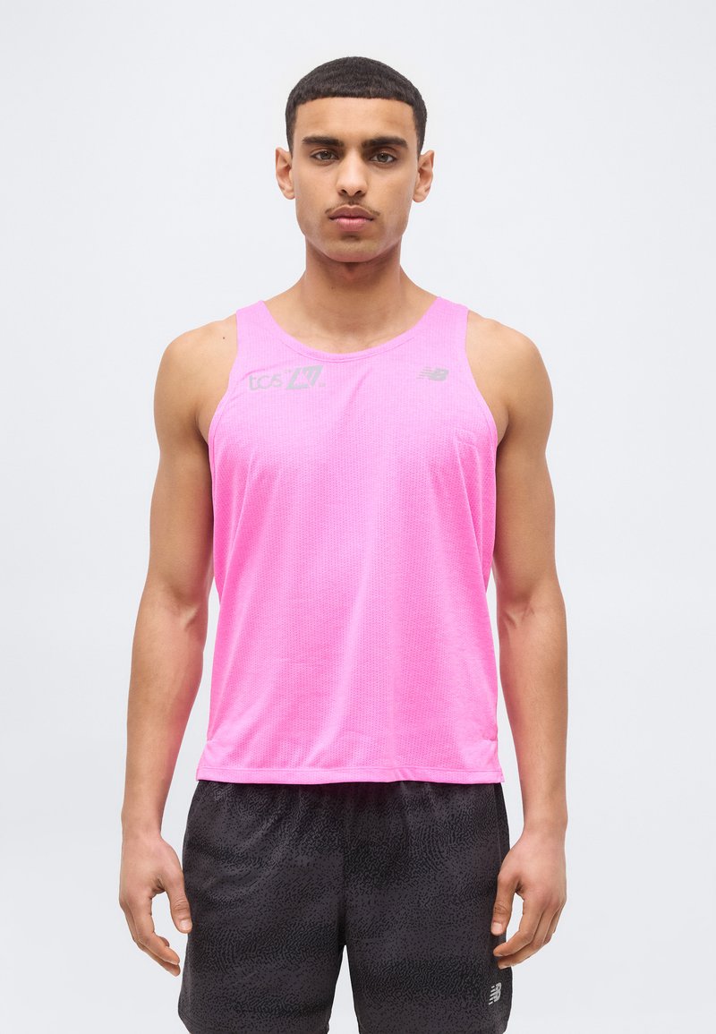 Νέος άνδρας στέκεται με το πρόσωπο προς τα εμπρός, φορώντας φούξια αμάνικη αθλητική μπλούζα και μαύρο σορτς με υφή, σε απλό φόντο.