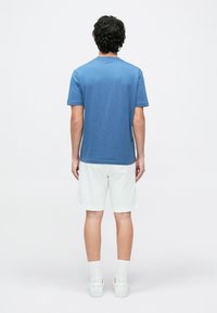 Mees seisab seljaga pärja, kandes sinist lühikeste varrukatega särki, valgeid lühikesi pükse, valgeid sokke ja valgeid tossusid tavalise tausta ees.