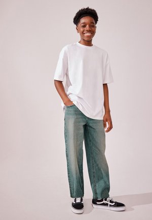 Jeune personne souriante debout, portant un t-shirt blanc oversize, un jean large et des baskets noir et blanc, une main dans la poche.