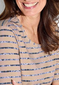 Beige gestreepte top met donkerblauwe en lichtblauwe ongelijkmatige strepen; ronde halslijn en een zachte stoftextuur. Haar dat langs de schouder valt.