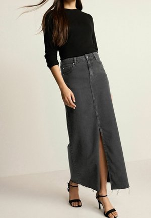 Kvinde iført sort bluse med lange ærmer, høj talje, falmet sort denim maxinederdel med frontsplit og sorte hæle med ankelrem foran en neutral baggrund.