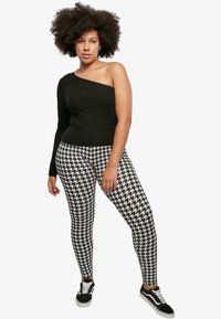 Fekete bordázott egyvállas felső fekete-fehér houndstooth leggingsszel párosítva, fekete sportcipővel viselve.