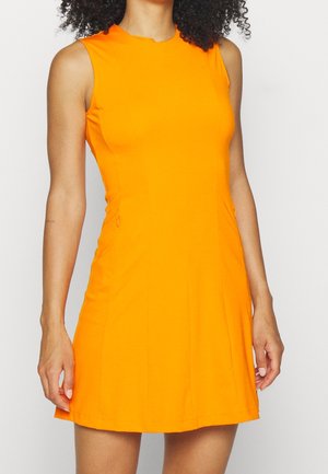 Kvinde med krøllet hår iført en ærmeløs, knaldorange A-linje kjole, der står mod en hvid baggrund.