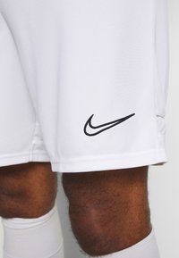 Primer plano de piernas de piel oscura que llevan pantalones cortos deportivos blancos con un logo negro de Nike y calcetines blancos hasta la rodilla.
