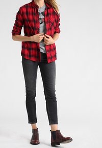 Camisa de franela a cuadros en rojo y negro con botones al frente, llevada sobre una camiseta gráfica gris, combinada con jeans skinny negros y botas de tobillo burdeos.