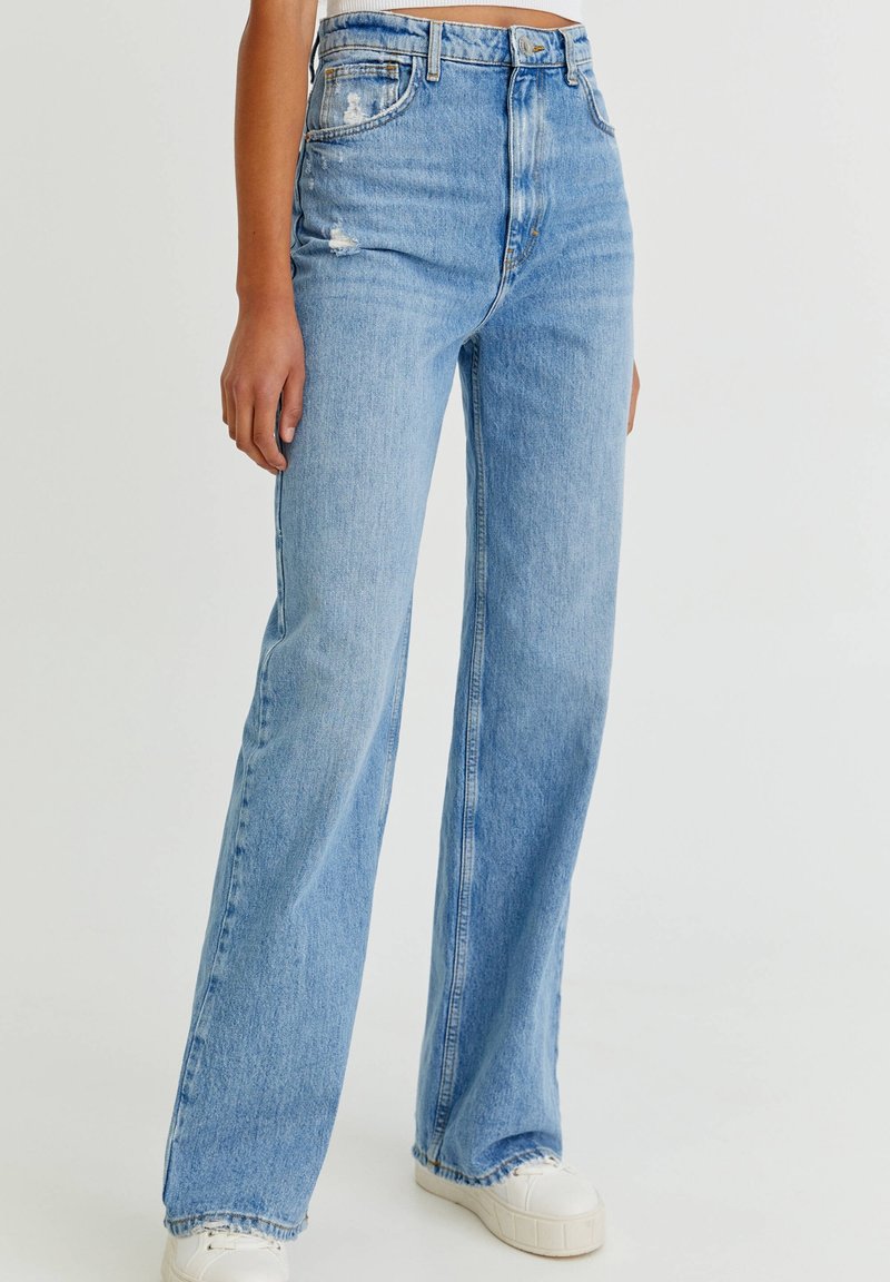 Jeans de denim azul claro con una cintura alta, pierna ancha y detalles desgastados. Presenta un clásico diseño de cinco bolsillos y un ligero desvanecimiento.