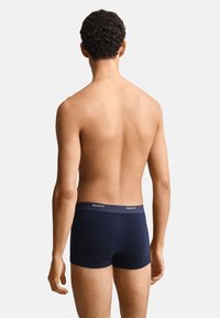 Homme debout, dos à la caméra, portant un boxer bleu marine avec une ceinture GANT sur fond blanc.