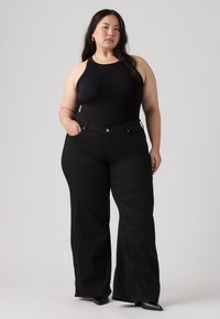 Γυναίκα με μαύρο αμάνικο τοπ, μαύρο παντελόνι με φαρδιά μπατζάκια και μαύρα μυτερά παπούτσια, στέκεται με το ένα χέρι στην τσέπη σε απλό φόντο.