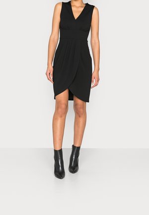 Woman wearing a sleeveless black wrap dress with a deep V-neck paired with black ankle boots, standing on a light grey background.