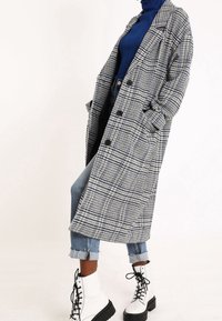 Longo cappotto grigio a scacchi con tasche profonde, bottoni neri e colletto strutturato, indossato sopra un dolcevita blu e jeans rovinati con risvolti.