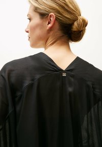 Indumento nero trasparente con scollo a V, caratterizzato da un accento metallico decorativo sul retro e tessuto fluido e leggero.