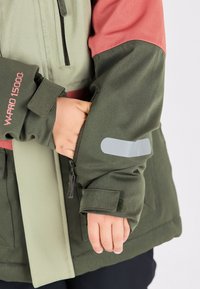 Chaqueta verde y gris claro con acentos rosas, que cuenta con un bolsillo con cremallera y un detalle reflectante en la manga. Tejido texturizado.