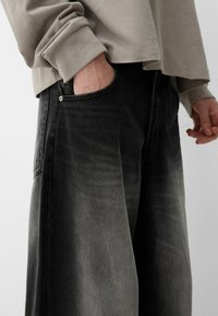 Farkkuhousut mustalla denim-kankaalla, joissa on haalistunut gradienttiefekti ja sivutaskut, metallinappiyksityiskohdalla varustettuna, yhdistettynä harmaaseen pitkähiharaiseen puseroon.