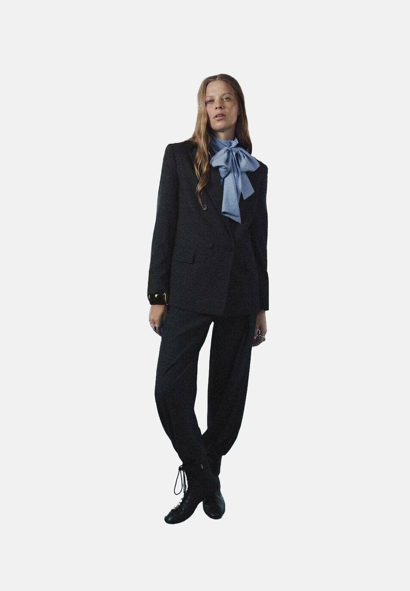 Blazer negro a medida con dos botones, combinado con pantalones negros de corte holgado. Un lazo azul claro agrega un toque de contraste. Zapatos negros de cordones.