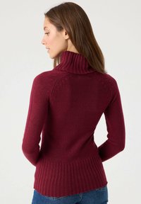 Suéter de punto burdeos con un alto cuello acanalado, silueta ajustada y dobladillo acanalado. Presenta un patrón texturizado a lo largo de los hombros.